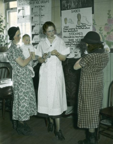 A health care worker is instructing two mothers how to select shoes and stockings for their children. On the wall is a poster on clothing and shoes.
