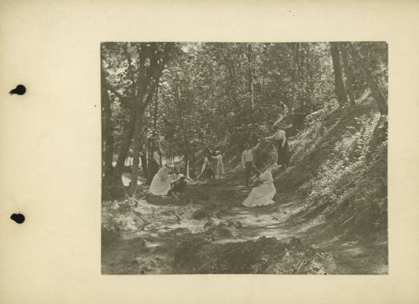 Page from photo album showing Nina S. Dousman and three sisters posed outdoors while hiking in a coulee, north of town. Nina is seated and reading. Three other men and women are posed with them.