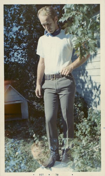 Candid portrait of Gordon Boardman, who is wearing a white shirt and a kerchief around his neck. There is a double exposure ghost image of him at the bottom of this image and behind him is a small doghouse.