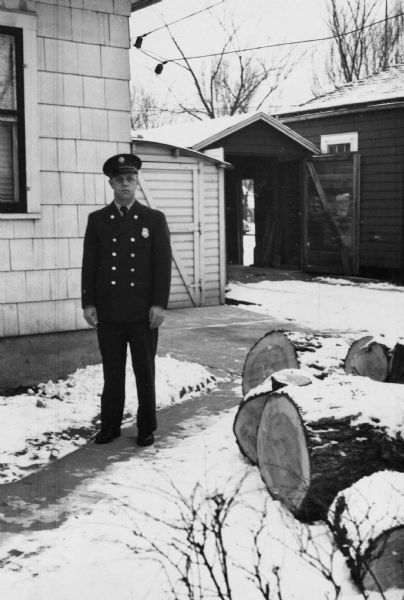 George William Piernot (1922-2000) standing outdoors wearing his Madison Fire Department uniform near the garage and home of his parents George Francis and Rhody (Boyum) Piernot at 709 North Street. Piernot served as a member of the Madison Fire Department from about 1946 until his retirement in 1977. He primarily worked at stations 7 and 9, where he drove the ladder truck.
