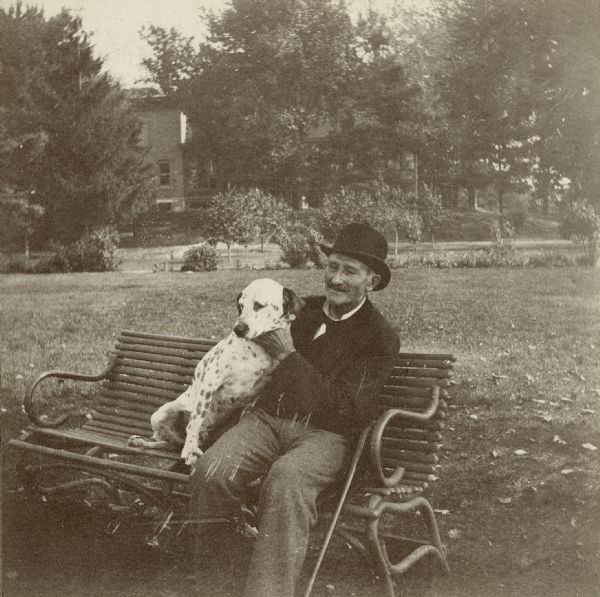 Man wearing a hat is seated on bench with a dalmatian dog.