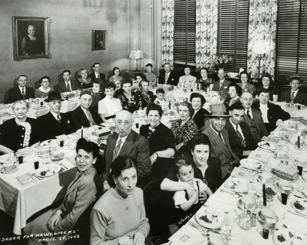 A group portrait of people seated at tables for Seder. The Pelz family if identified in the lower right of the picture. The mother is identified as Esther Pelz, holding her son Harry Pelz. The father, Nathan Pelz, is to her left and is wearing a hat.