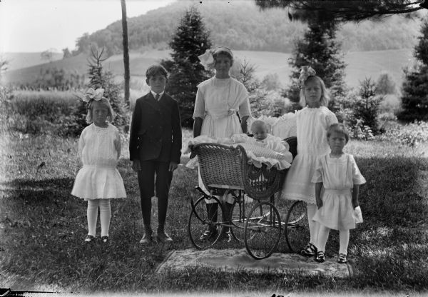 Group of six children posed outdoors for a group portrait. An infant is in a baby carriage, the other children are  standing.