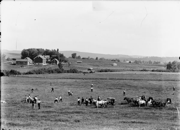 View from distance of group playing a game of baseball. Spectators sit in horse-drawn carriages. The game is being played in a farm field, with farm buildings and farmhouse in the background.