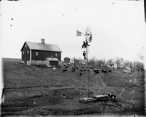 View up hill of a Woodmanse Mfg Co. windmill. Three men seated are posing near the top of the windmill, which stands in a field where cows are grazing. In the background is a barn and farm buildings. A group of people stands in and around a carriage near the barn.