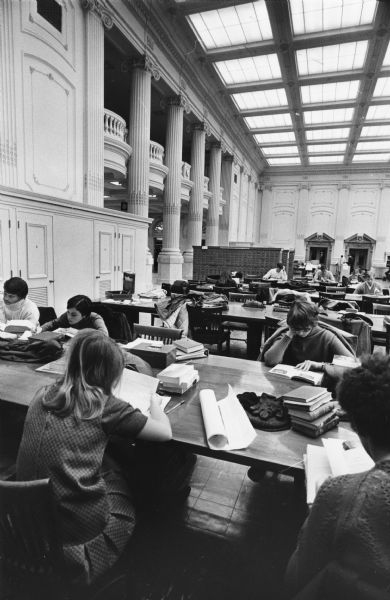 University students studying in the wisconsin Historical Society's library reading room. This view is undated, but the presence of the microfilm viewing rooms along the west wall suggests that it was taken before the remodeling of the mid-1960s when the separate microfilm reading room was created.