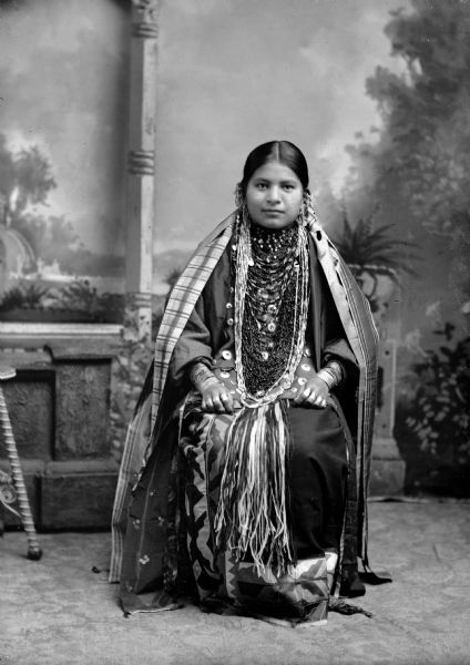 Studio portrait of a Ho-Chunk woman sitting and wearing several bead necklaces, earrings, and file bracelets. Her name is Emma Thunder, Littlesoldier (WePaMaKaRaWinKah) (Daughter of [WaConChaKah] John Thunder aka Dr. Thunder and [WeHonPeKaw] Lucy Bear, Thunder.) In the background is a painted backdrop.