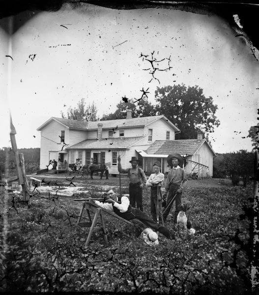 This is the second Herman Amberg Preus Spring Prairie parsonage.  Four men in the foreground pose with rakes and tools. One man sights along a shotgun. Other people pose with a carriage, and a group poses on the porch of frame house.