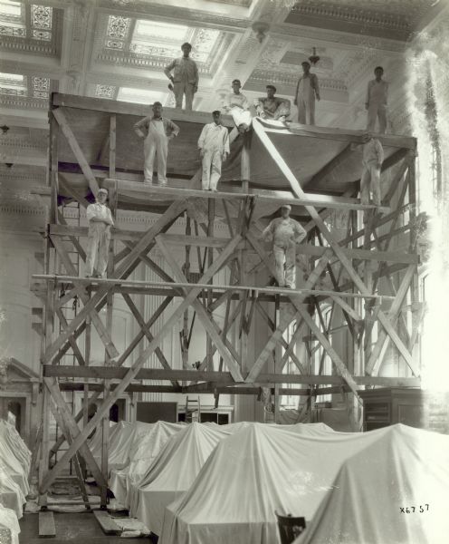 Men in work clothes stand on scaffolding in the library reading room of the Wisconsin Historical Society. Desks below are covered with drop cloths. The reading room skylight can be seen above the workers.