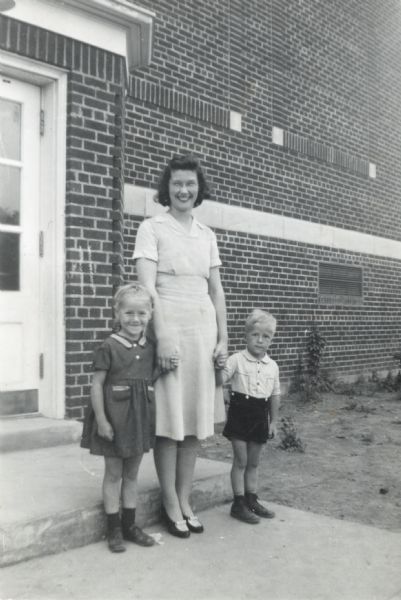A portrait of Winnifred Ahrens Spring holding hands with a young girl and a young boy, standing outside of the doorway of a building. Winnifred was a kindergarten teacher at West Side School in Rhinelander, Wisconsin.