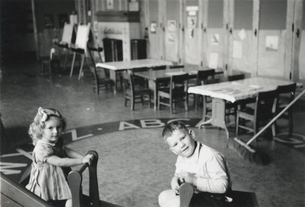 A young boy and a young girl playing in their kindergarten classroom. Desks, chairs, easels, a fireplace, and a broom are visible in the background.
