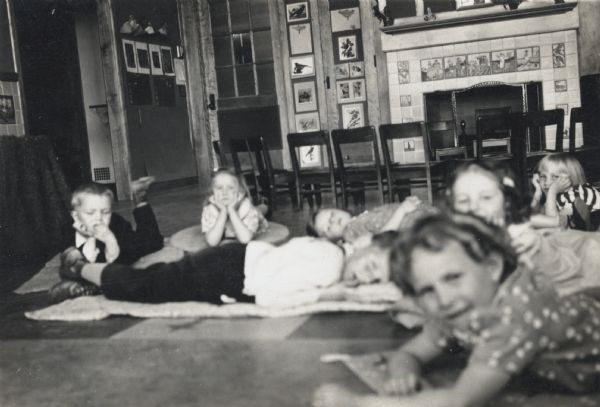 A group of students laying down on the floor of their kindergarten classroom, probably for nap time. In the background there is a fireplace surrounded with many chairs and lots of artwork on the walls. The fireplace is also surrounded by tiling depicting fairy tales and nursery rhymes.