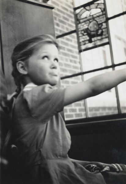A young girl posed near a window and glancing up. The window has a stained glass panel, possibly depicting the nursery rhyme character Humpty Dumpty.