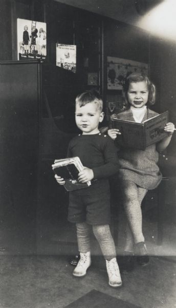 Two young students holding books in their kindergarten classroom next to a piano. The young girl is posed seated on a piano bench and holding an open book. The young boy is posed standing on the left and holding several closed books.