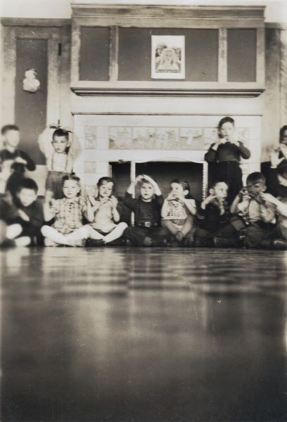 A group of kindergarten students posed sitting and standing near the fireplace in their classroom. The fireplace is surrounded by tiling depicting fairy tales and nursery rhymes.