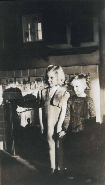 A portrait of two young students, a boy and a girl, standing next to each other and holding hands near the fireplace in their kindergarten classroom. The fireplace is surrounded by tiling depicting fairy tales and nursery rhymes.