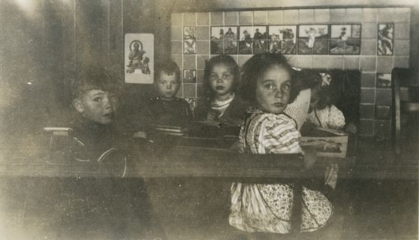 A group of five kindergarten students sitting around a table doing work near the fireplace in their classroom. The fireplace is surrounded by tiling depicting fairy tales and nursery rhymes.