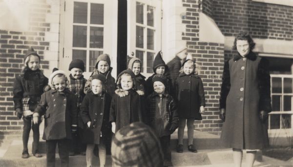 A group portrait of eleven kindergarten students and their teacher, Winnifred Ahrens Spring, standing outside of a building, probably the West Side School.