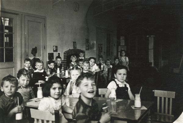 A view of young students at Lincoln School sitting around tables and drinking milk out of glass bottles. Behind the students in the background a fireplace is visible.