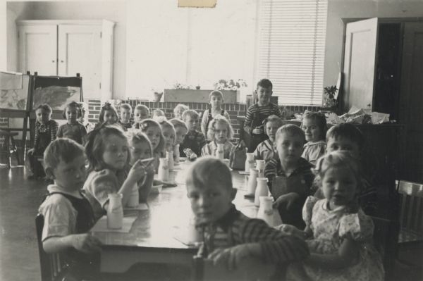 A view of young students at Lincoln School clustered around a long table and drinking milk out of glass bottles. In the background two art easels and plants on the windowsill are visible.

Winnifred Ahrens Spring introduced the free milk program to the Lincoln school.