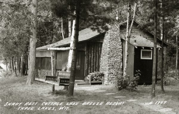 Exterior view of Shady Rest Cottage at Lake Breeze Resort. The building has a stone chimney and there is a wooden bench swing in the front yard. Caption reads: "Shady Rest Cottage, Lake Breeze Resort, Three Lakes, Wis."