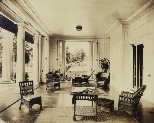 View of the west porch of Wadsworth Hall with wicker tables and chairs. There are papers on the table in the foreground and houseplants against the far wall.