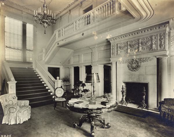 Interior view of the main hall of Wadsworth Hall. There is a table in the center of the room with books and a lamp, a fireplace at right and a stairway at left. A chandelier hangs above the base of the stairs.