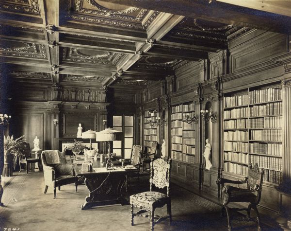 Interior view of the library at Wadsworth Hall. There is one large table with two lamps and several mismatched chairs. Sculptures and houseplants also decorate the room.