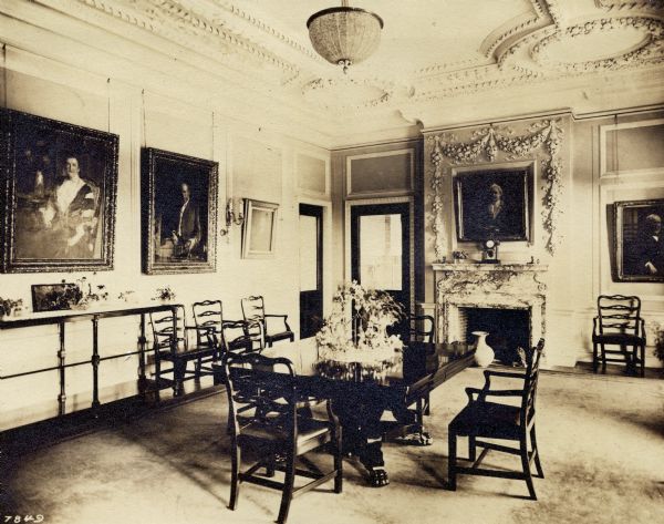 View of the dining room at Wadsworth Hall. There is a table with a centerpiece and four chairs. Other chairs line the walls of the room. There is a fireplace on the far wall and portraits on the two visible walls.