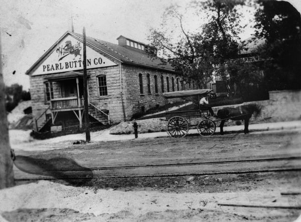 Exterior view from across road of the Pearl Button Company. A man sits in a horse and buggy parked outside the building.