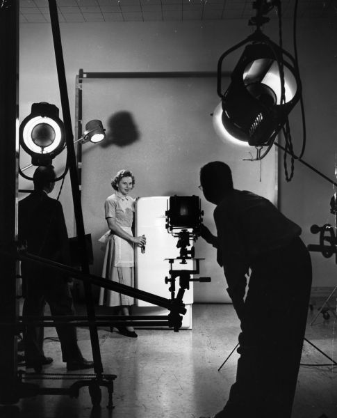 A woman wearing an apron posing with a refrigerator in a photography studio while a photographer takes a picture for advertising.