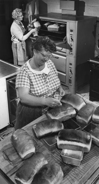 Elevated view of two women, one pulling loaves of bread from the oven and the other glazing crusts in the church kitchen.
