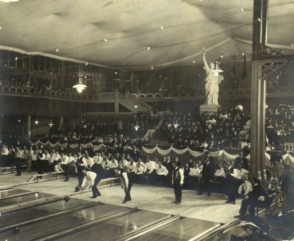 An audience watches bowlers at Milwaukee Auditorium. There is a Statue of Liberty replica in the seating area.