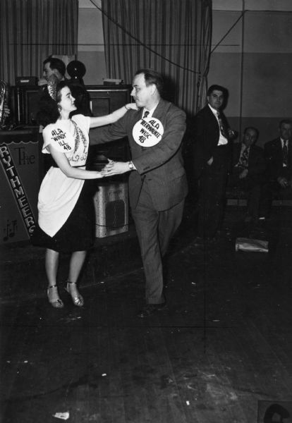 A man and a woman dance together at the Lithographers Intercity Bowling Dinner Dance. She is wearing a large tag that says "ALA 'Windy' Ill. 48" and he is wearing a tag that says "ALA 'Beerwaukee' Wisc. 48."