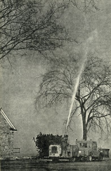 Worker spraying Elm trees with DDT pumped from a tank hitched to the back of a vehicle equipped with a snow plow.