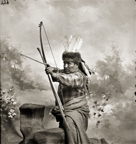 Studio portrait in front of a painted backdrop of a Ho-Chunk man, Hoonch-Schad-E-Gab (Big Bear), aiming a bow and arrow and wearing a fur hat with feathers.