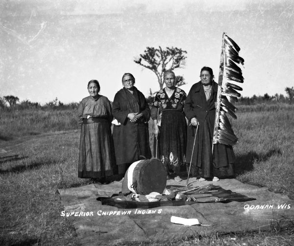 Four Odanah Ojibwa women standing outdoors at the edge of a blanket laying on the ground. The blanket has various ceremonial Ojibwa artifacts including a drum, drum sticks, and rattles. All the women are wearing long dresses, and one dress has a beaded yoke and hem. One woman is holding the women's veteran Eagle Staff that was made c. 1870. This eagle staff was damaged in a fire in the mid-1900's, but as of 2010 it still survives. Each eagle feather on the staff represents a deceased veteran.