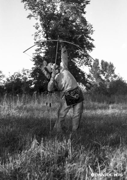 Ojibwa man shooting an arrow at the sky. The man is wearing buckskin leggings, a beaded apron, and porcupine roach. Two arrows are stuck in the ground beside him.