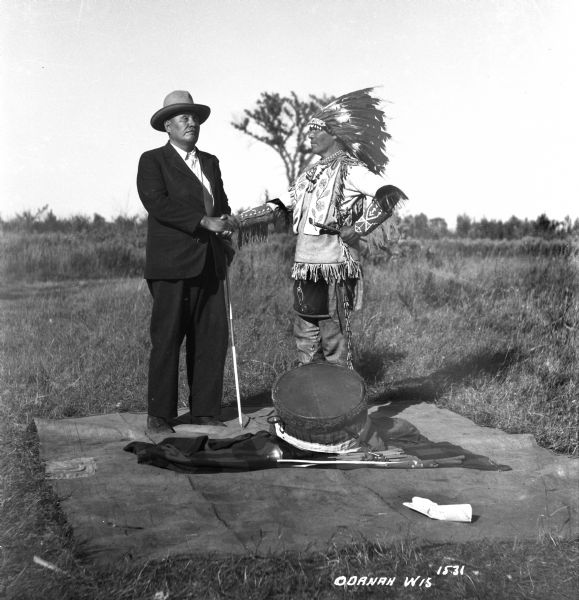 Two Ojibwa men standing outdoors on a blanket while shaking hands. The man on the left is dressed in a suit and wearing a hat while holding a blind walking cane. The other man is dressed in buckskin leggings, a beaded buckskin yoke, cloth arm bands, and wearing a feather war bonnet and holding drumsticks. A drum and drumsticks are on the blanket.