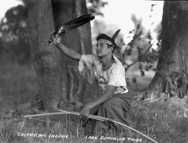 Ojibwa man kneeling on the ground, holding a bow and arrow in one hand and an eagle fan raised in the other hand. He is wearing a beaded headband.