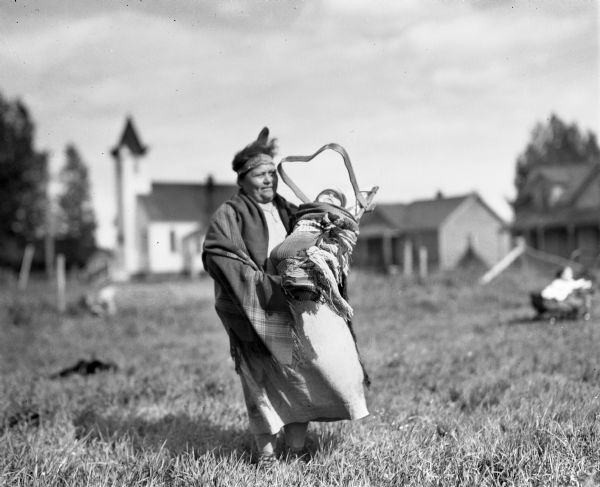 Ojibwa woman holding a baby in a tikinagan outdoors. The woman is wearing a dress, shawl and a headband. The Odanah Methodist church is in the background.