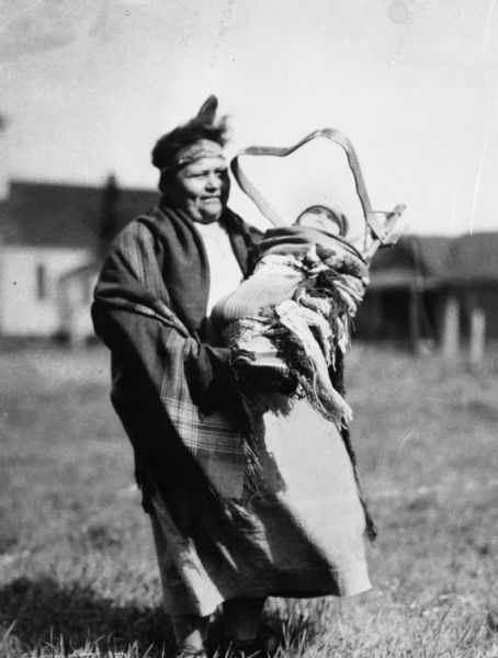 Bad River Ojibwe woman standing outdoors holding a baby in a tikinagan. The woman is wearing a dress, a shawl and a headband.  The Odanah Methodist church is in the background.