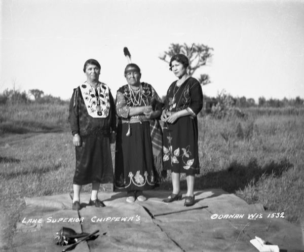 Three Bad River Ojibwa women standing outdoors on a blanket. The women are all wearing dresses with beadwork. The woman in the middle is wearing a headband with an eagle feather. There are drumsticks and a rattle on the blanket.