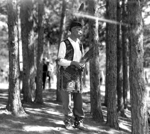 An Ojibwa man standing in a forest holding an eagle feather fan. His vest and apron are beaded and appliqued. He is wearing leather moccasins, ankle bells, and a porcupine roach.