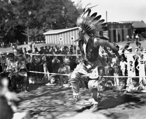 An Ojibwa man on a platform dancing for spectators. The dancer is wearing a feather war bonnet, leather vest, arm band, fringed buckskin leggings, moccasins, and ankle bells. Two Ojibwe children are sitting in the corner of the ring.