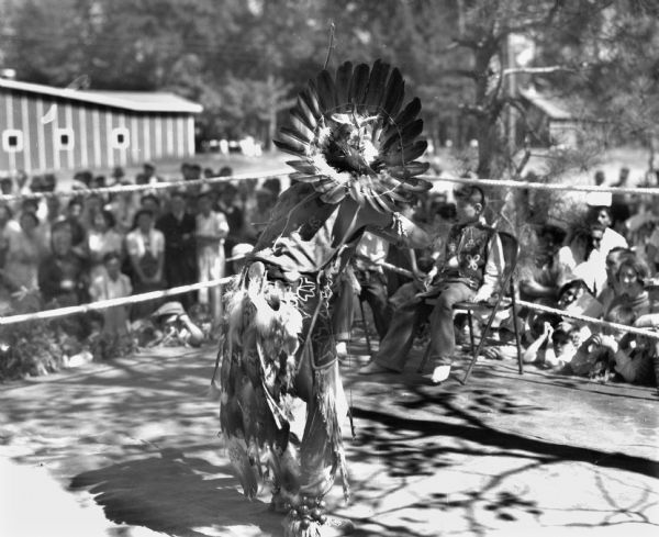 An Ojibwa man in on a stage dancing for spectators. The man is wearing a feather war bonnet, a beaded apron, leather moccasins, and ankle bells. An Ojibwa boy is sitting at the edge of the ring.
