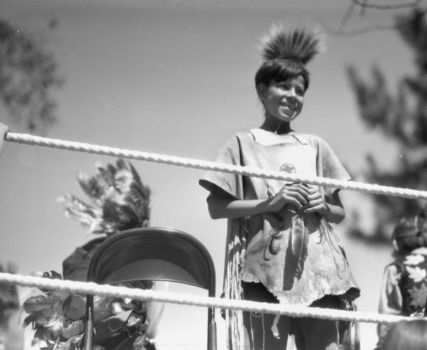 A smiling Ojibwa boy standing in a boxing ring holding an eagle feather. The boy is wearing a buckskin shirt with fringe and a porcupine roach.