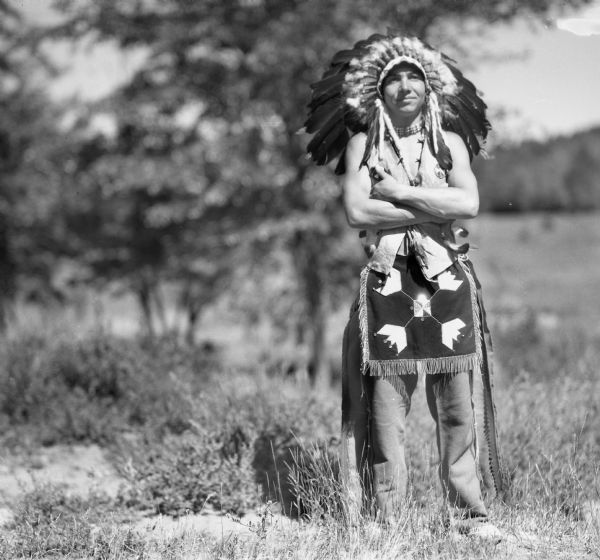 A smiling Ojibwa man standing in a field with his arms crossed. He is wearing an appliqued apron with fringe and beads over buckskin leggings, a beaded choker, and a feather war bonnet.