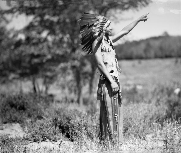 An Ojibwa man pointing toward the sky. He is wearing a beaded and appliqued apron over leggings, a buckskin vest, and a feather war bonnet.