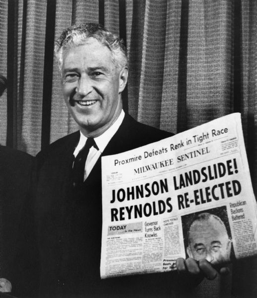 A smiling Warren Knowles displays a newspaper with the headline "Johnson Landslide! Reynolds Re-elected". Reynolds was Knowles' political opponent.